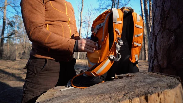 テーマハイキングと旅行。白人の観光男性は、オレンジ色のバックパックを解凍し、彼のものを取り出し、森の中の切り株にそれらを置きます。キャンプのための機器ともの — ストック写真