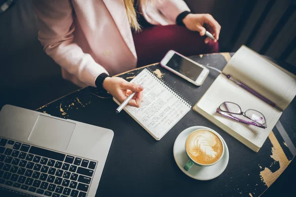 Close-up Kaukasische vrouw hand op houten tafel in cafe maakt notities in notebook. Onderwerp freelancer blogger journalist aan het werk. Onherkenbaar persoon. Technologie telefoon en laptop met kopje koffie — Stockfoto