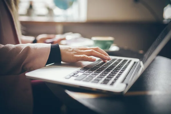 Vista superior de cerca de las manos de la mujer caucásica Estudiante, bloguero, escritor vestido casual que trabaja en una computadora portátil con un teléfono en la mano, dentro de la cafetería una mesa de madera y una taza de café — Foto de Stock