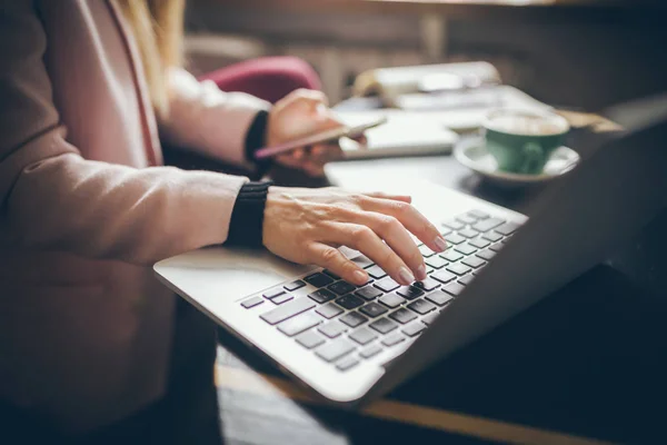 Κοντινό-up top θέα της λευκής γυναίκας χέρια περιστασιακά ντυμένος φοιτητής, blogger, συγγραφέας άνθρωπος που εργάζονται σε ένα φορητό υπολογιστή κρατώντας ένα τηλέφωνο στο χέρι του, μέσα στο καφέ ένα ξύλινο τραπέζι και ένα φλιτζάνι καφέ — Φωτογραφία Αρχείου