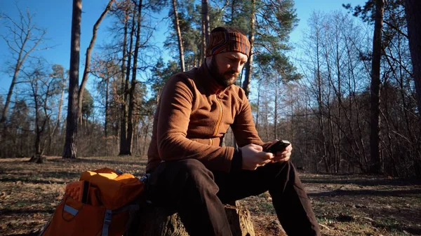Turystyka przyrodnicze podróży. Kaukaski mężczyzna turysta z brodą i plecak odpoczynku, Break odwrócił wyblakła drzewo, pniaka jesienią lasu sosnowego w słonecznej pogodzie. Wykorzystuje technologię ekranu dotykowego smartfona — Zdjęcie stockowe