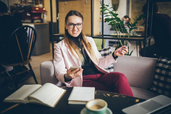 Tema profissão moderna blogueiro feminino. Mulher caucasiana com óculos e jaqueta sentada dentro da cafetaria atrás da mesa de madeira com notebook, laptop e xícara de café. Menina sonho emoção, pensar escritor — Fotografia de Stock