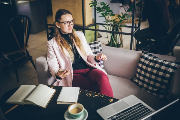 Tema profissão moderna blogueiro feminino. Mulher caucasiana com óculos e jaqueta sentada dentro da cafetaria atrás da mesa de madeira com notebook, laptop e xícara de café. Menina sonho emoção, pensar escritor — Fotografia de Stock