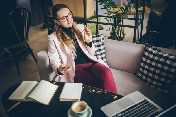 Tema profesión moderna blogger femenina. Mujer caucásica con gafas y chaqueta sentado dentro de la cafetería detrás de la mesa de madera con portátil, portátil y café taza. Chica emoción sueño, pensar escritor — Foto de Stock