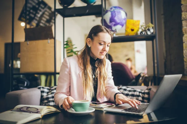 Mooie jonge Kaukasische vrouw het drinken van koffie en het typen op een toetsenbord in een cafe aan een houten tafel. Het onderwerp van de moderne beroepen is een blogger, freelancer en schrijver — Stockfoto