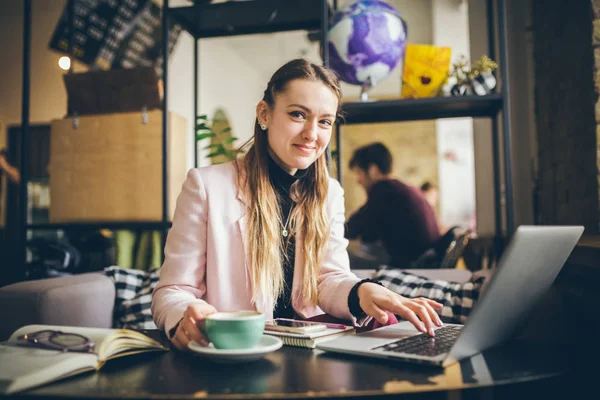 Mulher branca bonita bebendo café e digitando em um teclado dentro de um café em uma mesa de madeira. O tema das profissões modernas é um blogueiro, freelancer e escritor — Fotografia de Stock