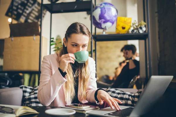 Beautiful young caucasian woman drinking coffee and typing on a keyboard inside a cafe at a wooden table. The topic of modern professions is a blogger, freelancer and writer