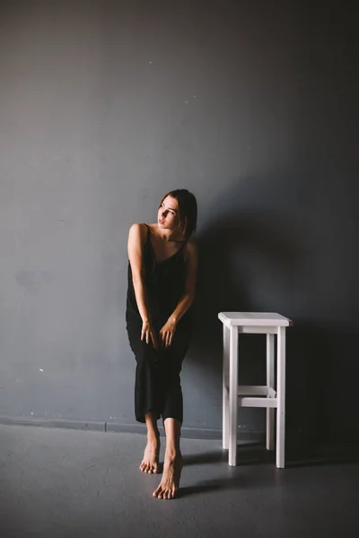 Tema social de la soledad femenina es el sufrimiento del dolor. Abastración de la violencia doméstica masculina. Joven hermosa mujer caucásica en ropa negra, pose extraña, silla blanca habitación oscura grande. Miedo y desesperación —  Fotos de Stock