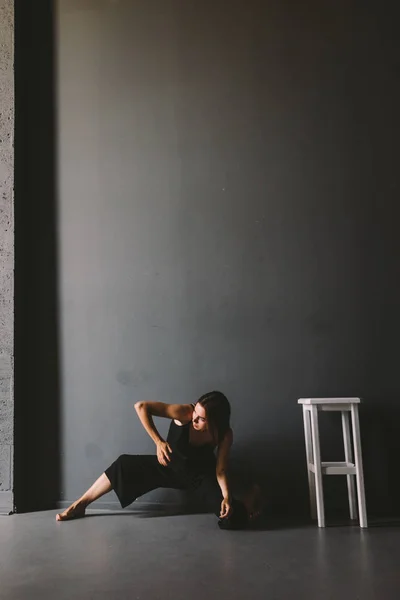 Social theme of female loneliness is pain suffering. Abastration of male domestic violence. Young beautiful Caucasian woman in black clothes, strange pose, white chair big dark room. Fear and despair — Stock Photo, Image