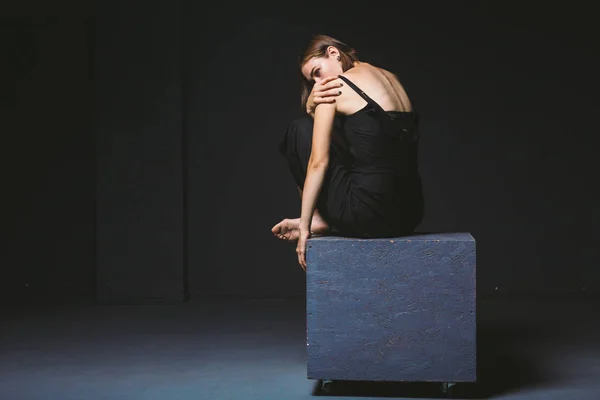 Thème social. jeune femme caucasienne en pose étrange en robe posant sur cube noir, fond sombre, symbolise la douleur, la souffrance, la recherche d'aide, la protection d'une société maléfique. Art moderne, abstraction — Photo