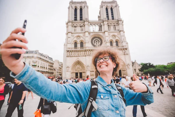 Tourist in Paris making funny selfie near Notre Dame Cathedral. Beautiful young Caucasian tourist woman with backpack in Paris making funny selfie hand holding phone, photo near Notre Dame Cathedral