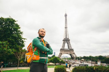 Elinde fotoğraf makinesi ve turuncu sırt çantası olan beyaz bir adam Fransa 'da duruyor. Paris, Eyfel Kulesi' nin arka planında.
