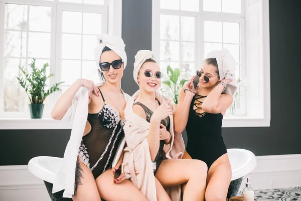 Theme female friendship. Party girlfriends, relaxation and spa procedures, hen party. Three Caucasian beautiful women sitting inside on bathroom in sunglasses, head towel and bathrobe, underwear