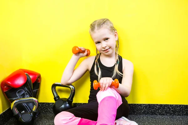 Deportes temáticos y salud de los niños. Niña divertida niña caucásica con coletas, se sienta descansando descanso en el suelo en el gimnasio. Equipos de mancuerna atleta para gimnasia culturismo fondo amarillo pared — Foto de Stock