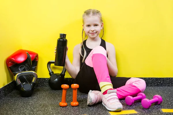 Theme sports and health children. Little Caucasian girl sits resting break floor gym holds hand bottle, drink water thirst. Athlete dumbbell equipments gymnastics bodybuilding background yellow wall