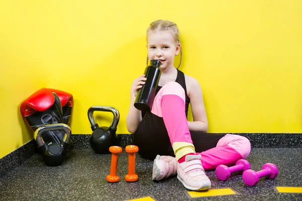 Deportes temáticos y salud de los niños. Niña caucásica se sienta descansando descanso gimnasio del piso sostiene la botella de la mano, beber sed de agua. Atleta mancuerna equipos gimnasia culturismo fondo amarillo pared — Foto de Stock