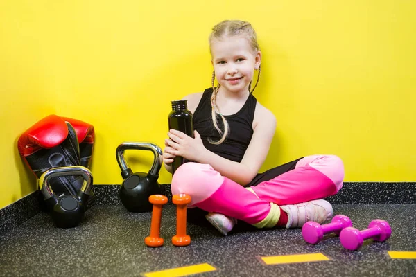 Deportes temáticos y salud de los niños. Niña caucásica se sienta descansando descanso gimnasio del piso sostiene la botella de la mano, beber sed de agua. Atleta mancuerna equipos gimnasia culturismo fondo amarillo pared —  Fotos de Stock