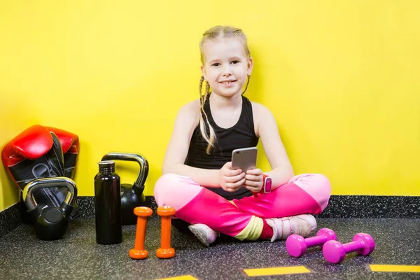 Young child girl with a mobile phone sitting on the floor near the dumbbells, boxing gloves and a bottle of water on the floor. Ready to workout at the gym. Sport and healthy concept