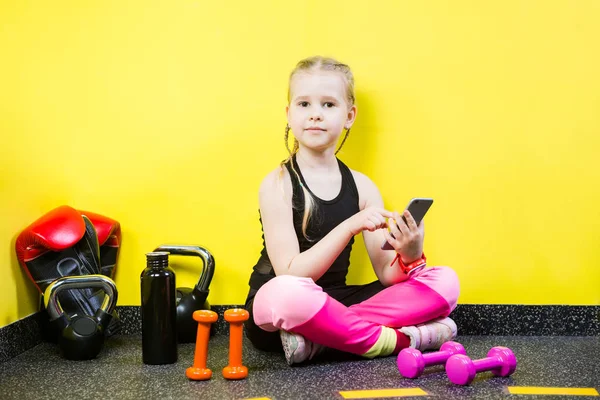 Niña pequeña con un teléfono móvil sentado en el suelo cerca de las mancuernas, guantes de boxeo y una botella de agua en el suelo. Listo para entrenar en el gimnasio. Deporte y concepto saludable — Foto de Stock