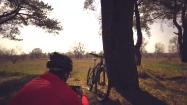 Genç beyaz kadın sporcu bisikletçi telefonla fotoğraf çekiyor, bisikletinin fotoğraflarını çekiyor, güneşli bir günde, doğa meydanındaki ağacın yanında duruyor. Miğferli ve spor giysili kız dinleniyor.