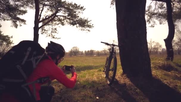 Jovem mulher branca atleta ciclista turístico faz foto no telefone, fotografa sua bicicleta de pé perto da árvore no campo da natureza dia ensolarado. Menina no capacete e sportswear descansando quebrar bicicleta de montanha — Vídeo de Stock