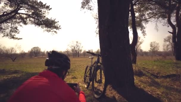 Jonge blanke vrouw atleet toerist fietser maakt foto 's op de telefoon, fotografeert zijn fiets staan in de buurt van boom op natuurgebied zonnige dag. Meisje in helm en sportkleding rustpauze mountainbike — Stockvideo