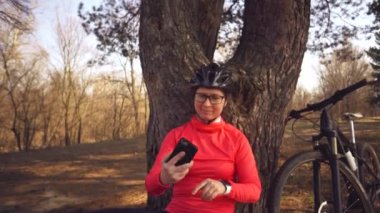 Beyaz kadın sporcu bisikletçi, kentin dışındaki kozalaklı ormandaki ağacın yanında otururken çekilmiş akıllı telefon fotoğrafını kullanıyor. Sporcu kadın dağ bisikletiyle selfie çekiyor.