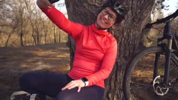 Young caucasian woman athlete tourist cyclist uses hand smart phone photo of herself selfie sitting near tree in coniferous forest outside the city. Sportswoman taking selfie with her mountain bike — 비디오