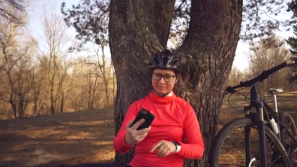 Молодая кавказская спортсменка-велосипедистка использует ручной смартфон, на котором она сидит рядом с деревом в хвойном лесу за городом. Спортсменка делает селфи на своем горном велосипеде — стоковое видео