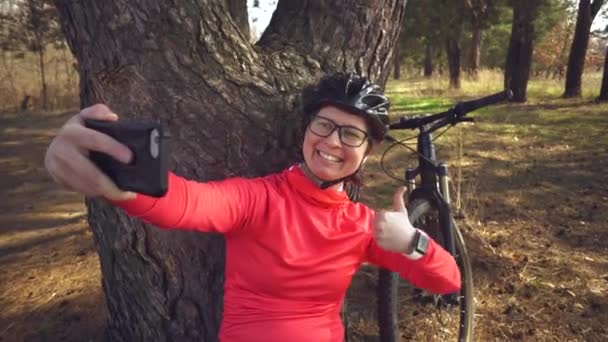 Joven atleta atleta caucásica ciclista turística utiliza la mano teléfono inteligente foto de sí misma selfie sentado cerca del árbol en el bosque de coníferas fuera de la ciudad. Deportiva tomando selfie con su bicicleta de montaña — Vídeos de Stock