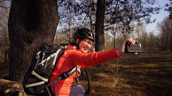 Young caucasian woman athlete tourist cyclist uses hand smart phone photo of herself selfie sitting near tree in coniferous forest outside the city. Sportswoman taking selfie with her mountain bike — Stock Photo, Image