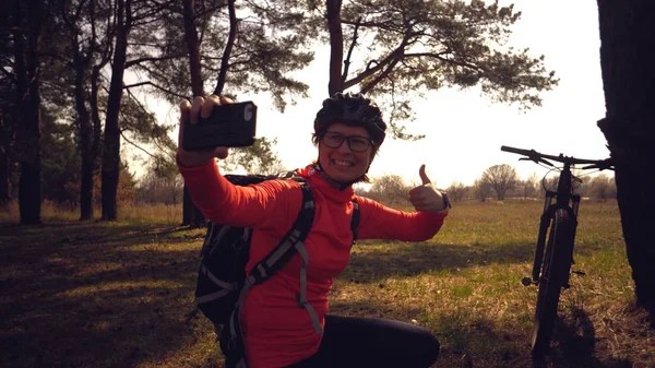 Joven atleta atleta caucásica ciclista turística utiliza la mano teléfono inteligente foto de sí misma selfie sentado cerca del árbol en el bosque de coníferas fuera de la ciudad. Deportiva tomando selfie con su bicicleta de montaña —  Fotos de Stock