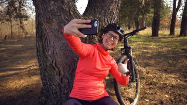 Young caucasian woman athlete tourist cyclist uses hand smart phone photo of herself selfie sitting near tree in coniferous forest outside the city. Sportswoman taking selfie with her mountain bike clipart