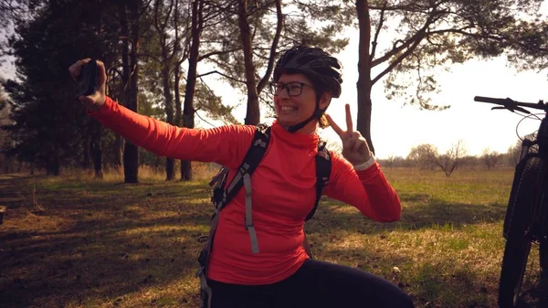Молодая кавказская спортсменка-велосипедистка использует ручной смартфон, на котором она сидит рядом с деревом в хвойном лесу за городом. Спортсменка делает селфи на своем горном велосипеде — стоковое фото