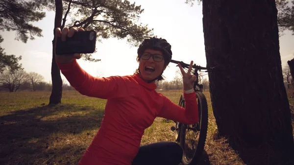 Joven atleta atleta caucásica ciclista turística utiliza la mano teléfono inteligente foto de sí misma selfie sentado cerca del árbol en el bosque de coníferas fuera de la ciudad. Deportiva tomando selfie con su bicicleta de montaña — Foto de Stock