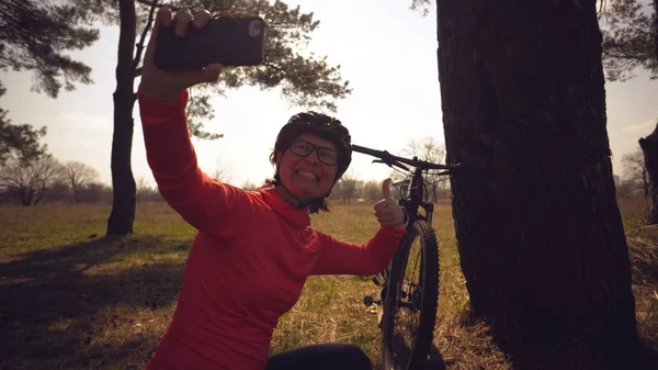 Joven atleta atleta caucásica ciclista turística utiliza la mano teléfono inteligente foto de sí misma selfie sentado cerca del árbol en el bosque de coníferas fuera de la ciudad. Deportiva tomando selfie con su bicicleta de montaña — Foto de Stock