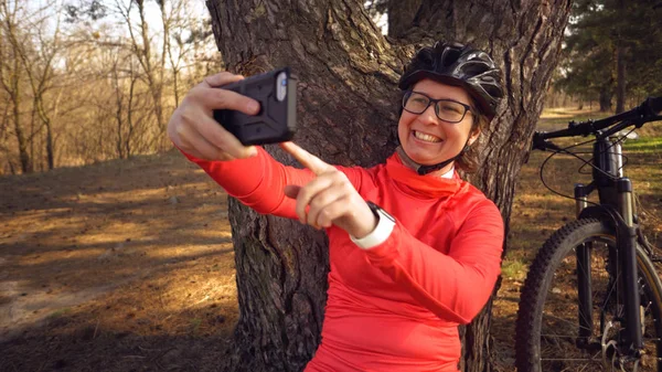 Молодая кавказская спортсменка-велосипедистка использует ручной смартфон, на котором она сидит рядом с деревом в хвойном лесу за городом. Спортсменка делает селфи на своем горном велосипеде — стоковое фото