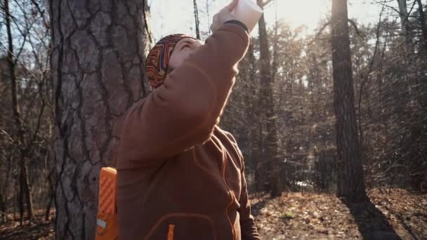 A young tired Caucasian man carries a rest break with a backpack sitting and drinking water on a tree in a nature forest during hikes, trips, vacations, on vacation, outdoor lifestyle freedom concept — Stock Video