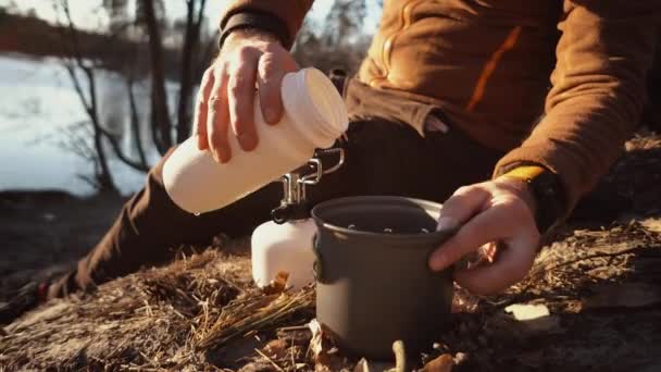 Mains homme Gros plan verse l'eau des flacons dans une casserole pour faire bouillir l'eau sur un brûleur à gaz touristique camping forêt — Video