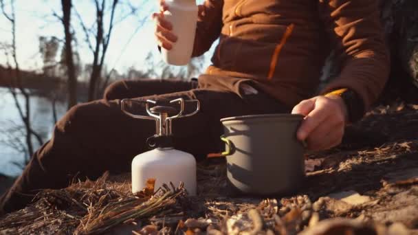 Mains homme Gros plan verse l'eau des flacons dans une casserole pour faire bouillir l'eau sur un brûleur à gaz touristique camping forêt — Video