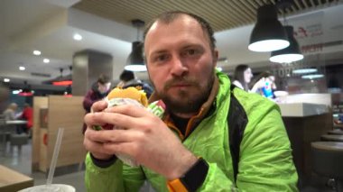 Sakallı ve spor giyimli beyaz adam akşam ları masadaki bir restoranın içinde hamburger ya da hamburger dolu abur cuburlarla ağzını doldurdu.