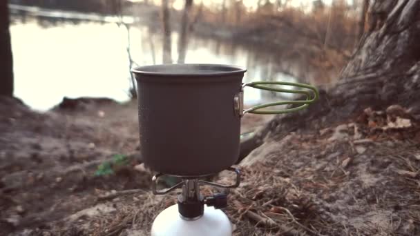 Zbliżenie palnika gazowego, na którym stoi czajnik z pokrywką, sprowadza wodę, parę wodną, gotowanie w warunkach zewnętrznych. To sprowadza jedzenie. Na brzegu rzeki w lesie. 4K — Wideo stockowe