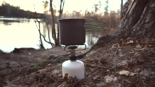 Close-up de um queimador de gás, em que uma chaleira com uma tampa está, ferve água, vapor, cozinhar em condições ao ar livre. Ferve comida. Na margem do rio na floresta. 4K — Vídeo de Stock