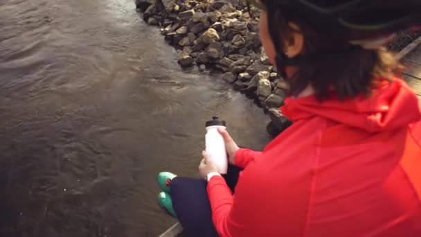 Parlak spor ayakkabı kask Kafkas kadın sporcu köprü üzerinde nehir çapraz dinleniyor. Elinde plastik bir şişe şişesi olan parlak spor giyimli bisikletçi. Dinlenme mola içki su susuzluk — Stok video