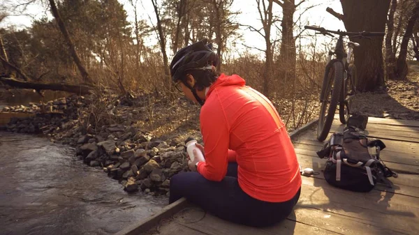 Καυκάσια γυναίκα αθλητής σε φωτεινά αθλητικά παπούτσια κράνος βρίσκεται στη γέφυρα διασχίζουν το ποτάμι. Ποδηλάτης σε φωτεινά αθλητικά είδη με ένα φλασκί ένα πλαστικό μπουκάλι στο χέρι του. Ανάπαυση διακοπή διάλειμμα ποτό νερό δίψα — Φωτογραφία Αρχείου