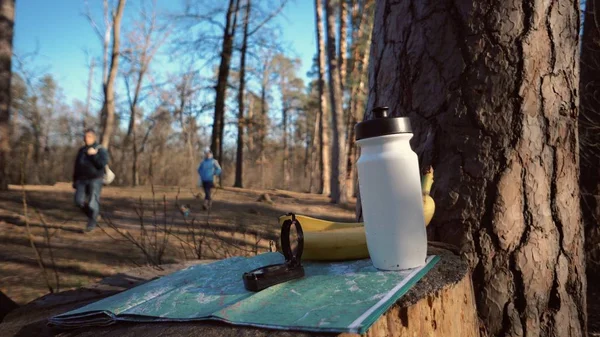 things for tourism travel for eco tourism in nature forest. Paper map, compass and snack food bottle flask with water and bananas fruit on stumped hemp tree. People walking blurred background