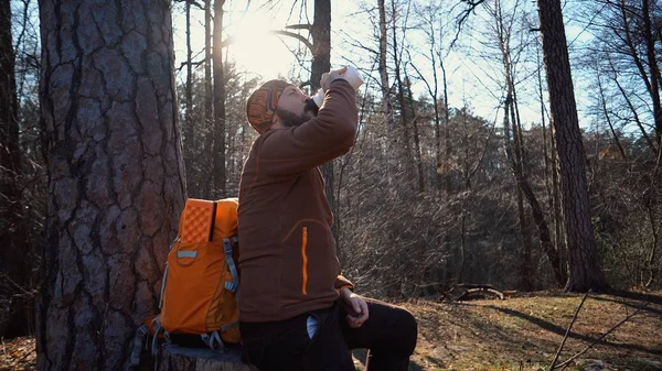 Ein junger müder kaukasischer Mann macht bei Wanderungen, Ausflügen, Urlauben, im Urlaub, im Freien eine Pause mit einem Rucksack und trinkt Wasser auf einem Baum im Naturwald — Stockfoto