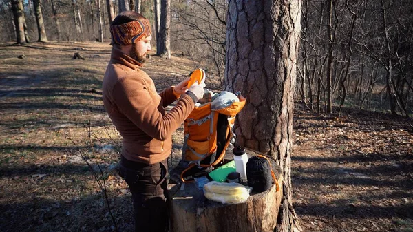Tema de senderismo y viajes. Un turista caucásico desempaca una mochila naranja, saca sus cosas y las pone en un tocón en el bosque. Equipamiento y cosas para acampar — Foto de Stock