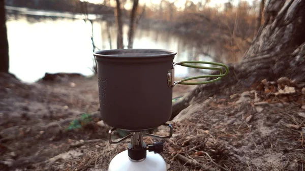 Κοντινό στάδιο ενός καυστήρα αερίου, στο οποίο ένας βραστήρας με καπάκι στέκεται, βράζει νερό, ατμός, μαγείρεμα σε εξωτερικές συνθήκες. Βράζει το φαγητό. Στην όχθη του ποταμού στο δάσος. 4K — Φωτογραφία Αρχείου