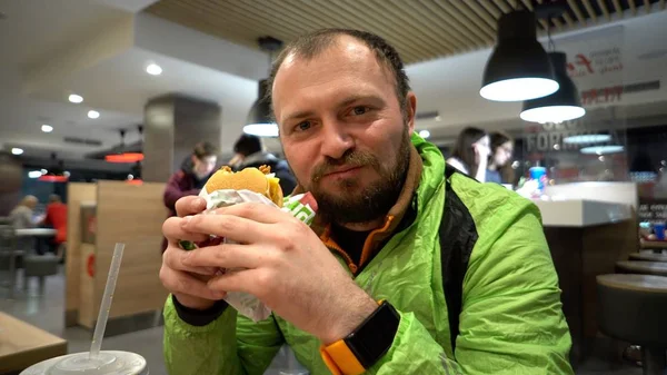 Kaukázusi Férfi a szakálla és a Sportruházat töltötte ki a száját a gyorsétel teljes egy hamburgert vagy hamburger belsejében egy étteremben az asztalnál az esti órákban — Stock Fotó